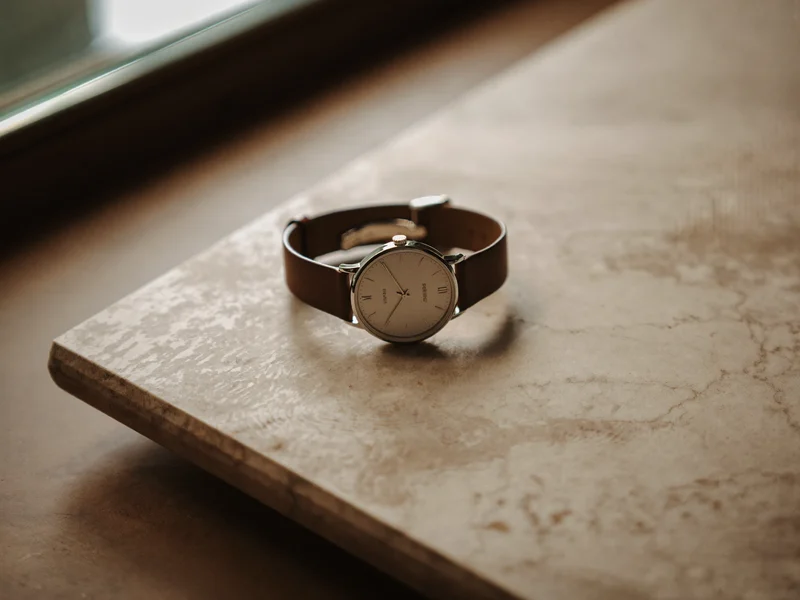 Schlanke Dresswatch mit unbeschriftetem Zifferblatt und braunem Lederband auf heller Steinfläche